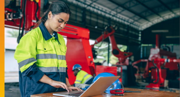 Engineer working on a laptop in a manufacturing workshop with industrial equipment in the background.